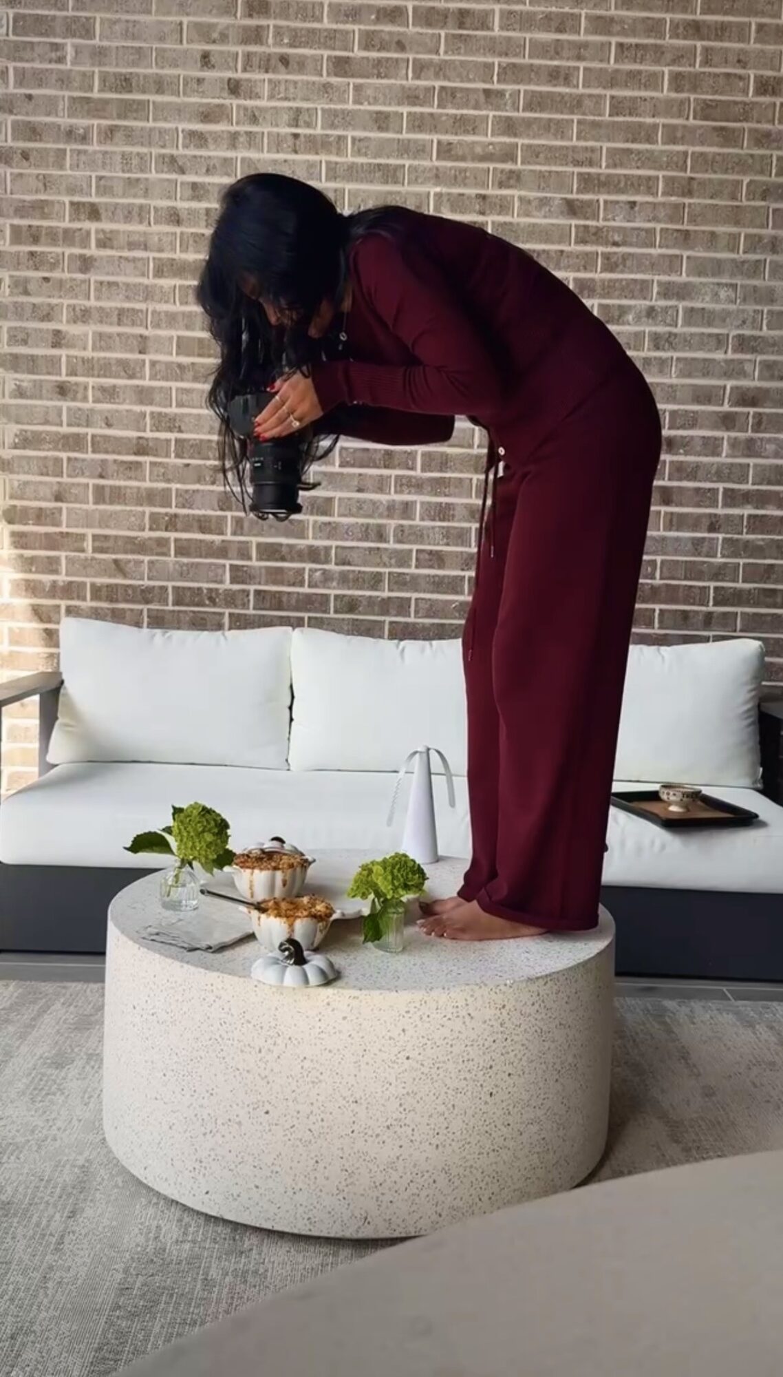 Person with dark hair in a maroon outfit stands on a round table, leaning forward, holding a camera, against a brick wall.