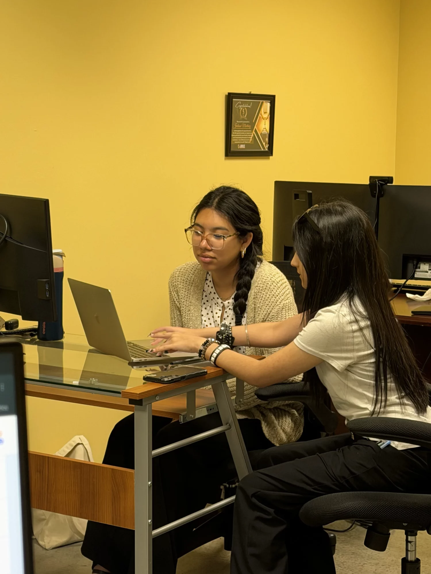 Two women sit at a desk with computers, one using a laptop, in a yellow-walled office.