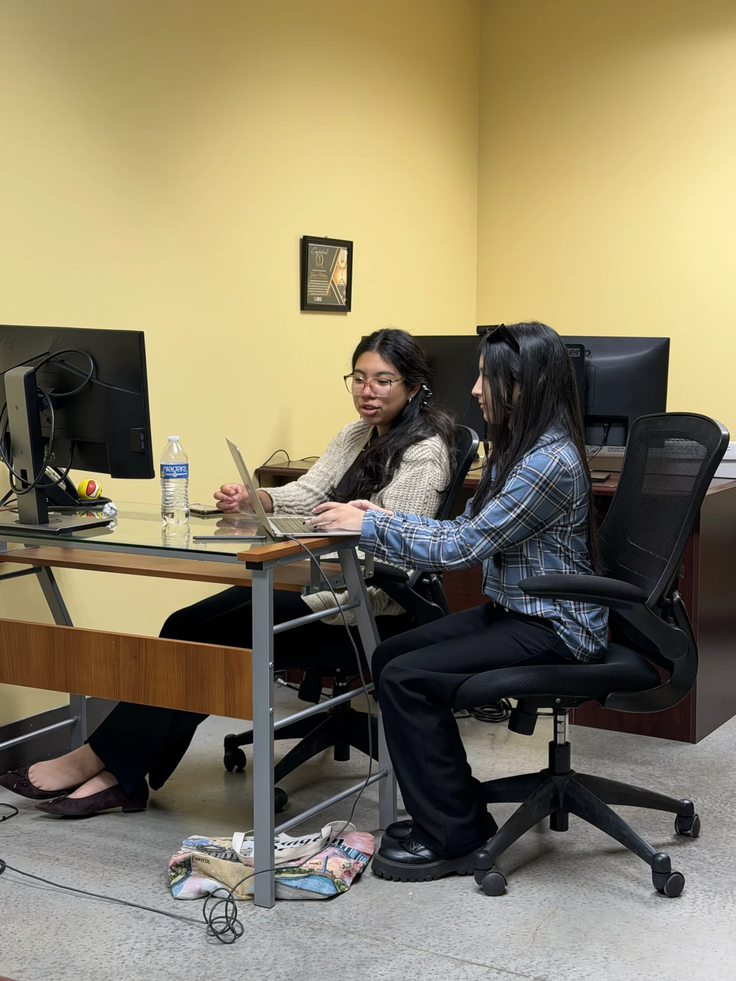 Two women sit at a desk in an office, looking at a phone together, with a computer, water bottle, and framed picture on the wall.