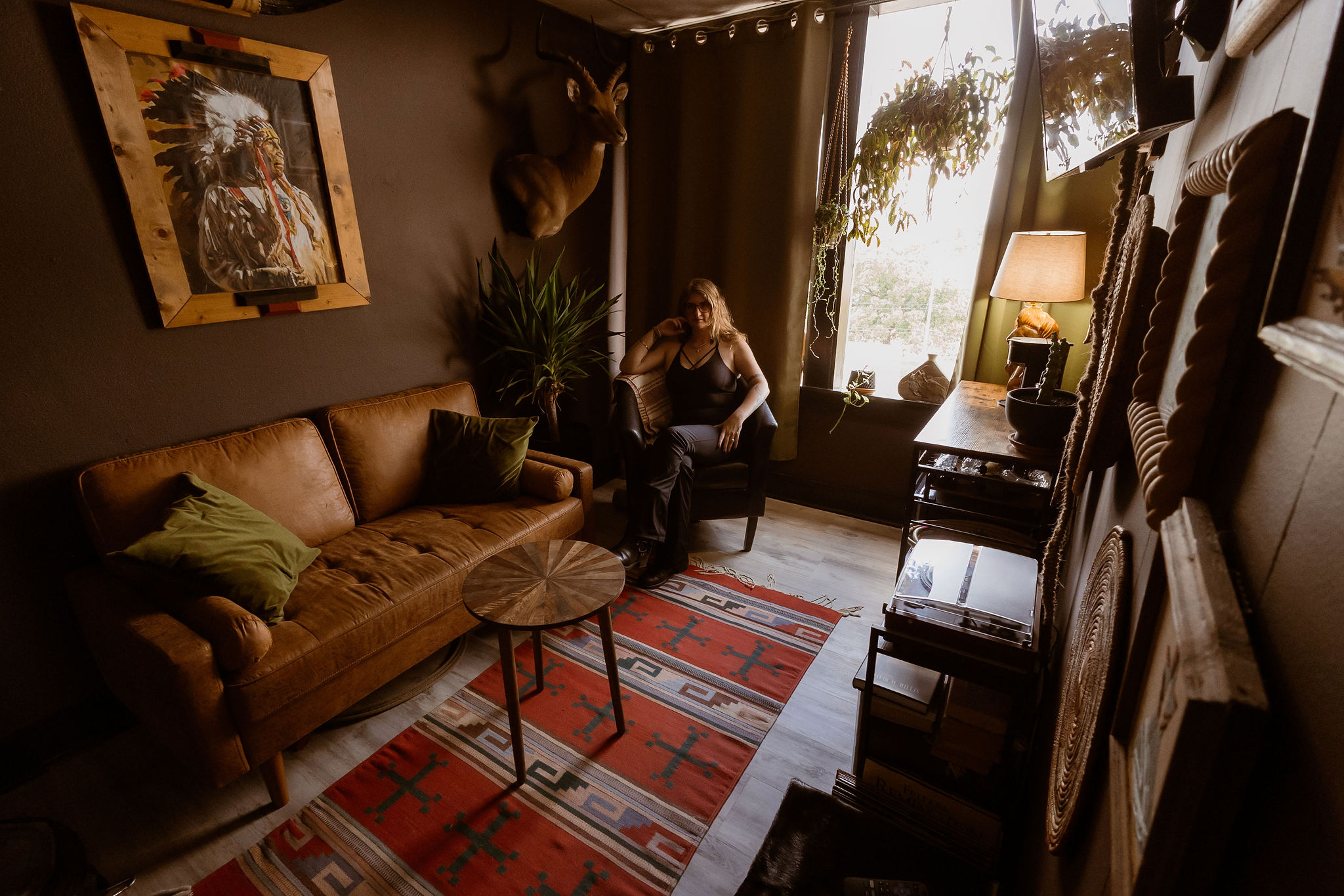 Living room with a woman sitting on a chair near a window, a brown sofa, a patterned rug, and a mounted deer head.