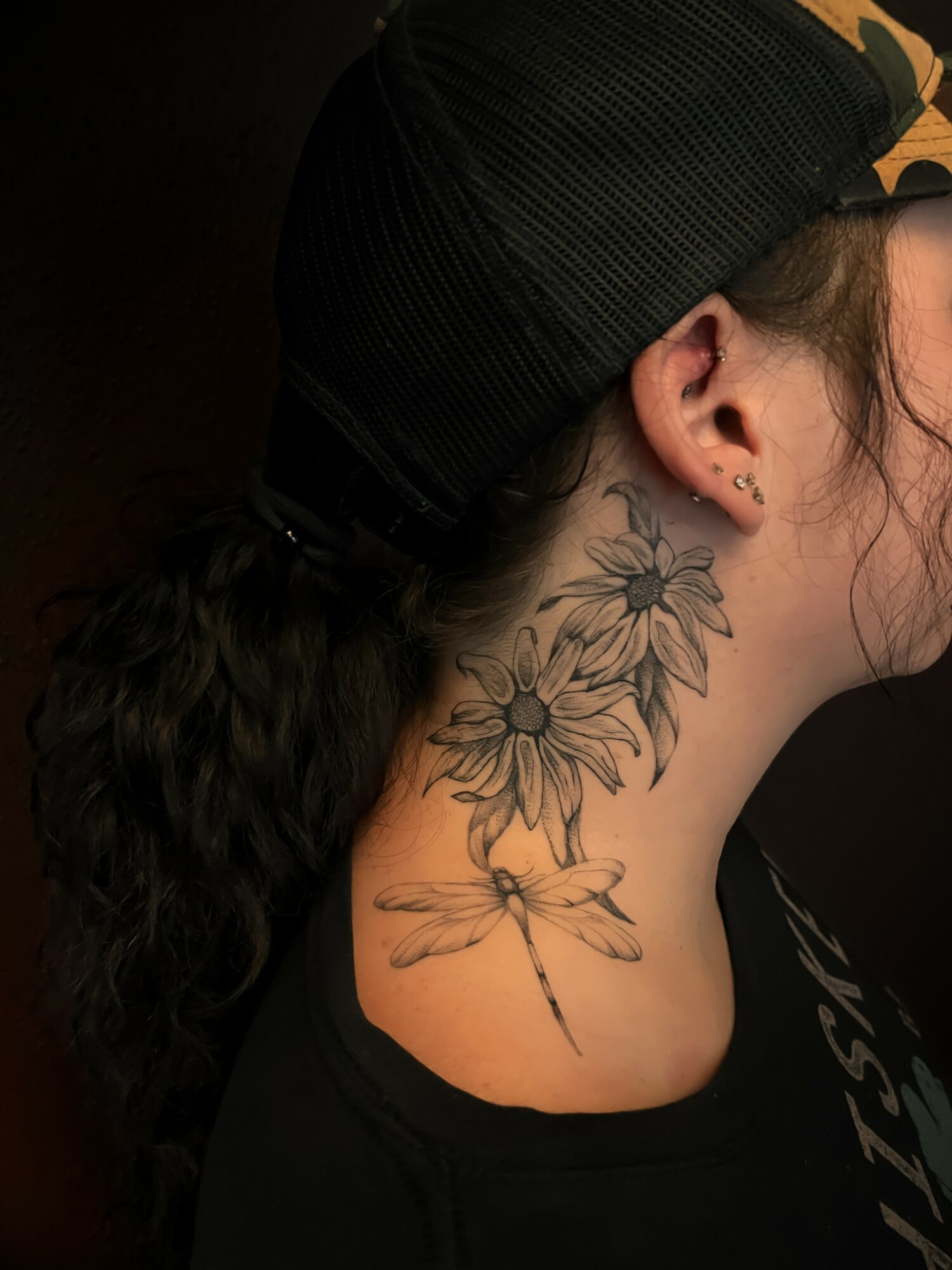 Side view of a person's neck with a floral and dragonfly tattoo, wearing a black cap and earrings.