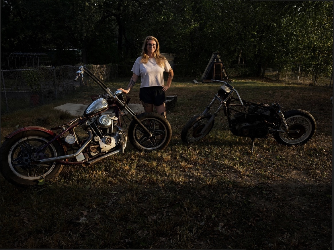 Woman standing between two motorcycles outdoors in a grassy area with trees in the background.