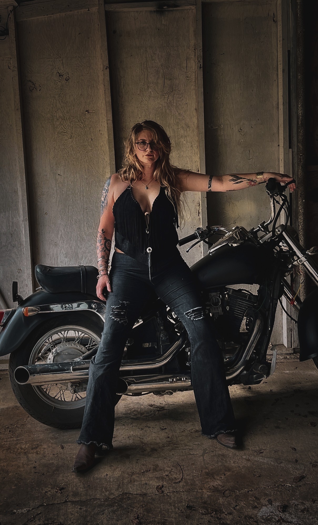 Woman with long hair and sunglasses standing next to a motorcycle in a concrete structure.