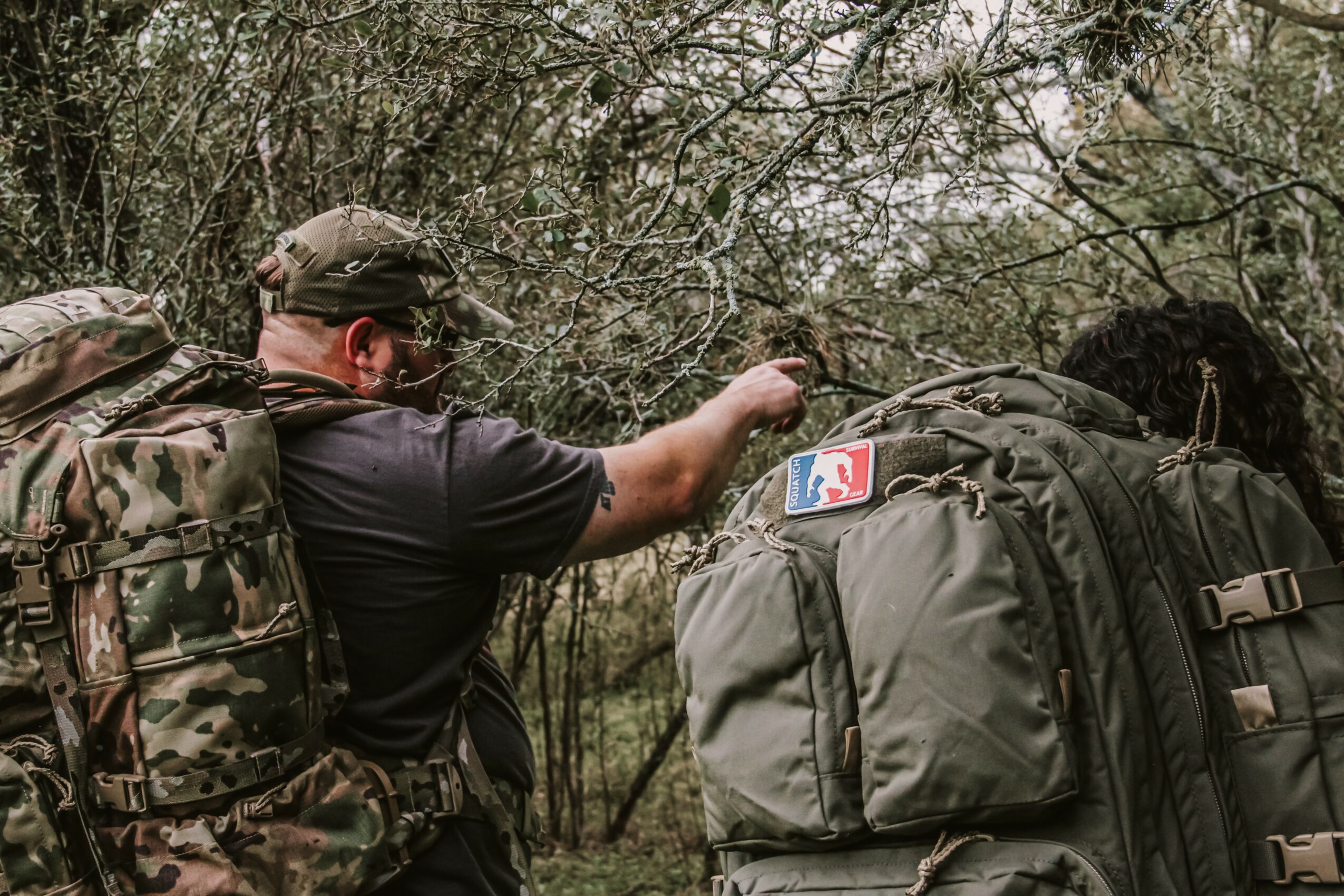 Person in camouflage gear with backpack reaching into wooded area, carrying a large backpack with a patch, outdoors.