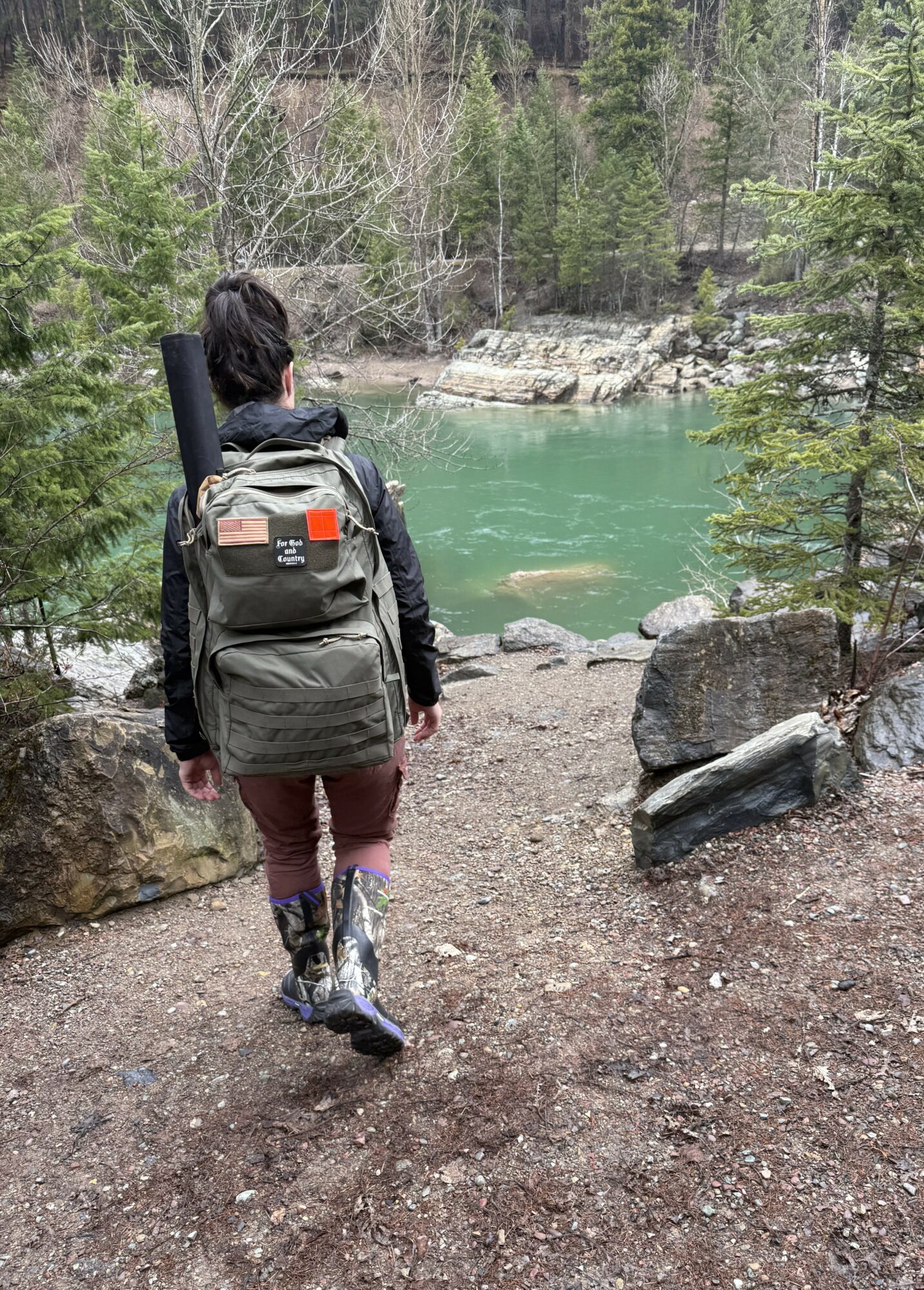 Person walking on a trail near a river surrounded by trees, carrying a backpack and a rolled mat, in a forested area.