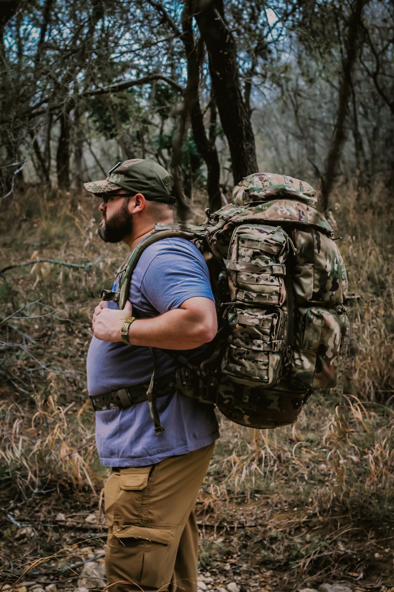 Man with a large backpack walking in a wooded area, wearing a cap, sunglasses, and casual outdoor clothing.