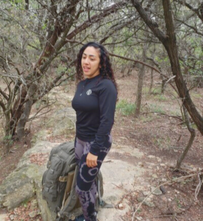 Woman standing outdoors among trees, holding a hat, with a backpack on the ground nearby.
