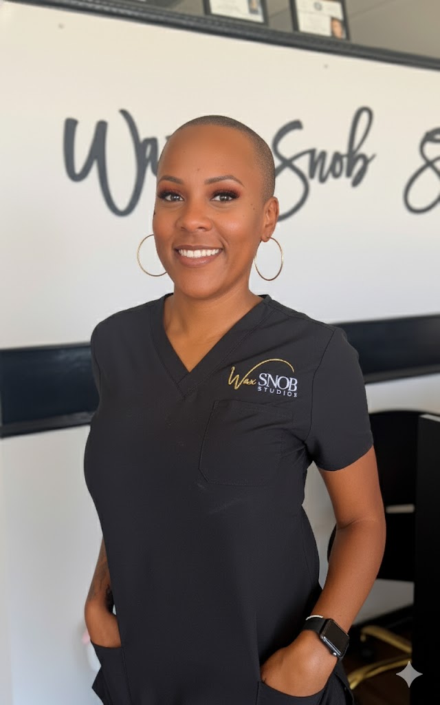 Smiling woman with a shaved head wearing black scrubs and hoop earrings, standing indoors.