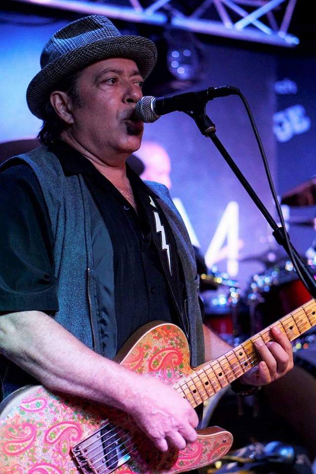 Man wearing a hat singing into a microphone while playing an electric guitar on stage.