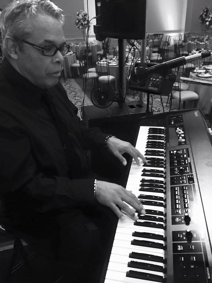 Person playing keyboard in a room with tables and chairs, microphone setup nearby, black and white photo.