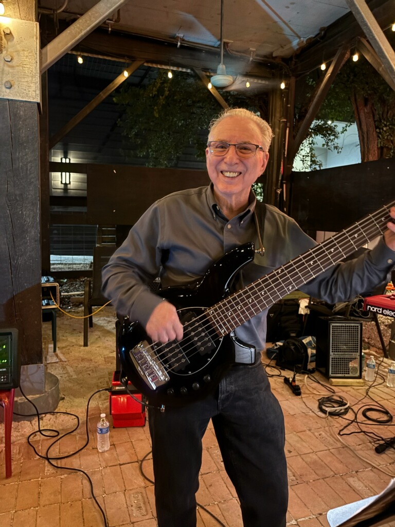Man smiling and playing a black electric bass guitar indoors, with musical equipment around him.