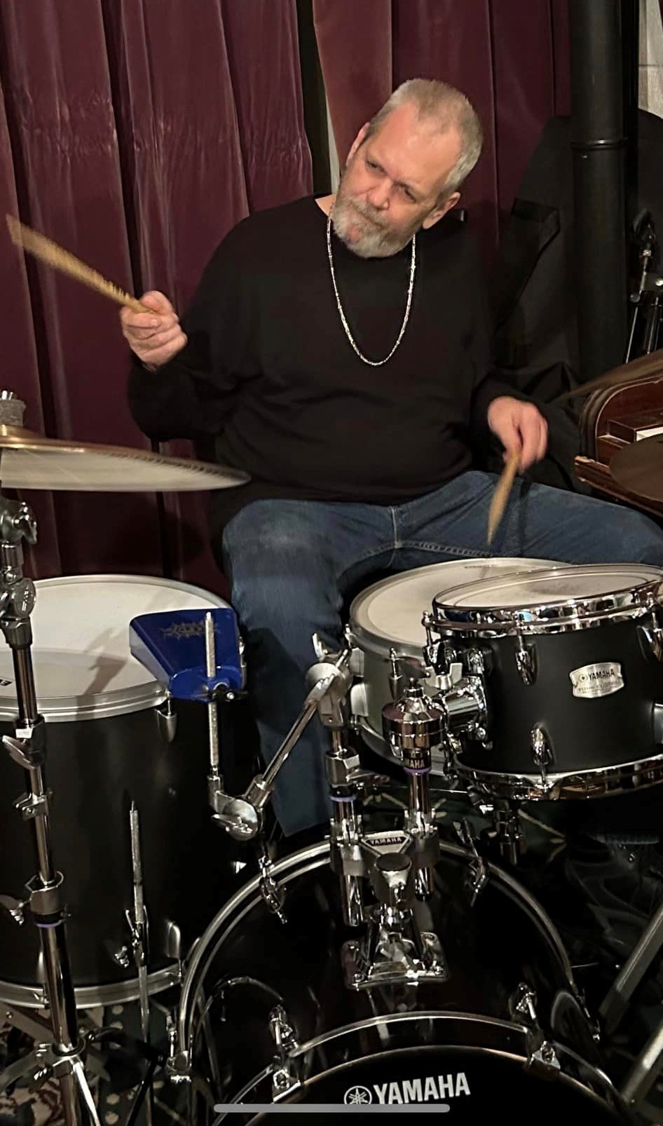 Man playing drums, holding drumsticks, wearing a black shirt and necklace, with a dark background.