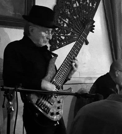 Person wearing a hat playing an electric bass guitar indoors, with a decorative fan and a person in the background.