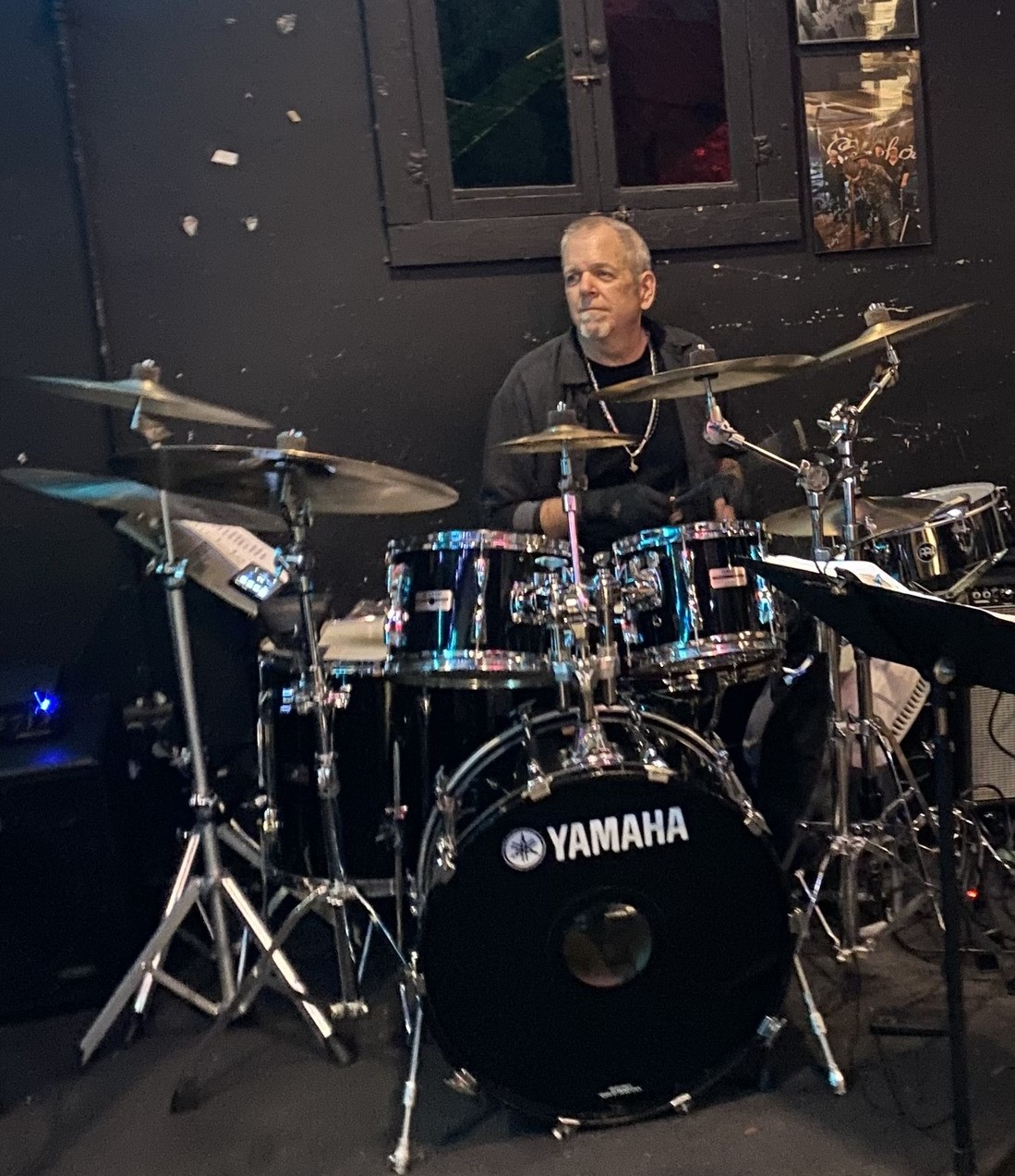 Man playing a Yamaha drum set in a dark room with black walls and window, surrounded by cymbals and drumsticks.