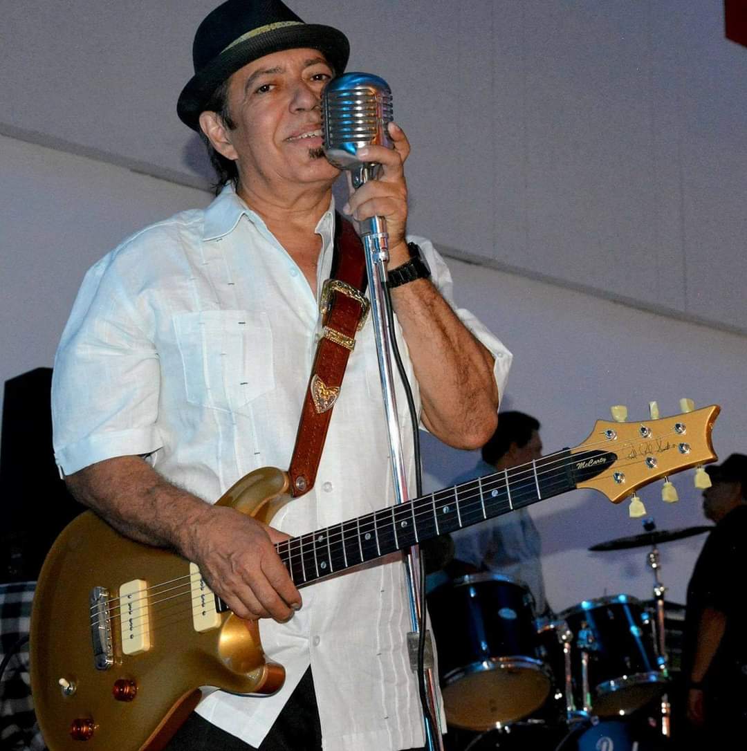 Man with a guitar singing into a vintage microphone, wearing a white shirt and a hat, with drums in the background.