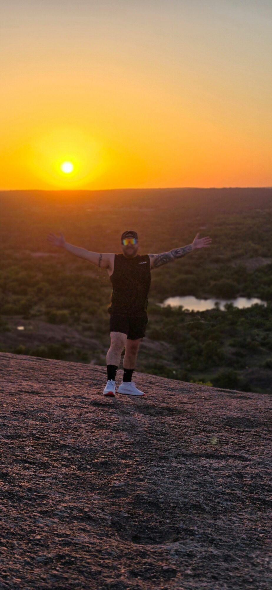 Person standing outdoors during sunset with a bright sun near the horizon, landscape and sky visible.