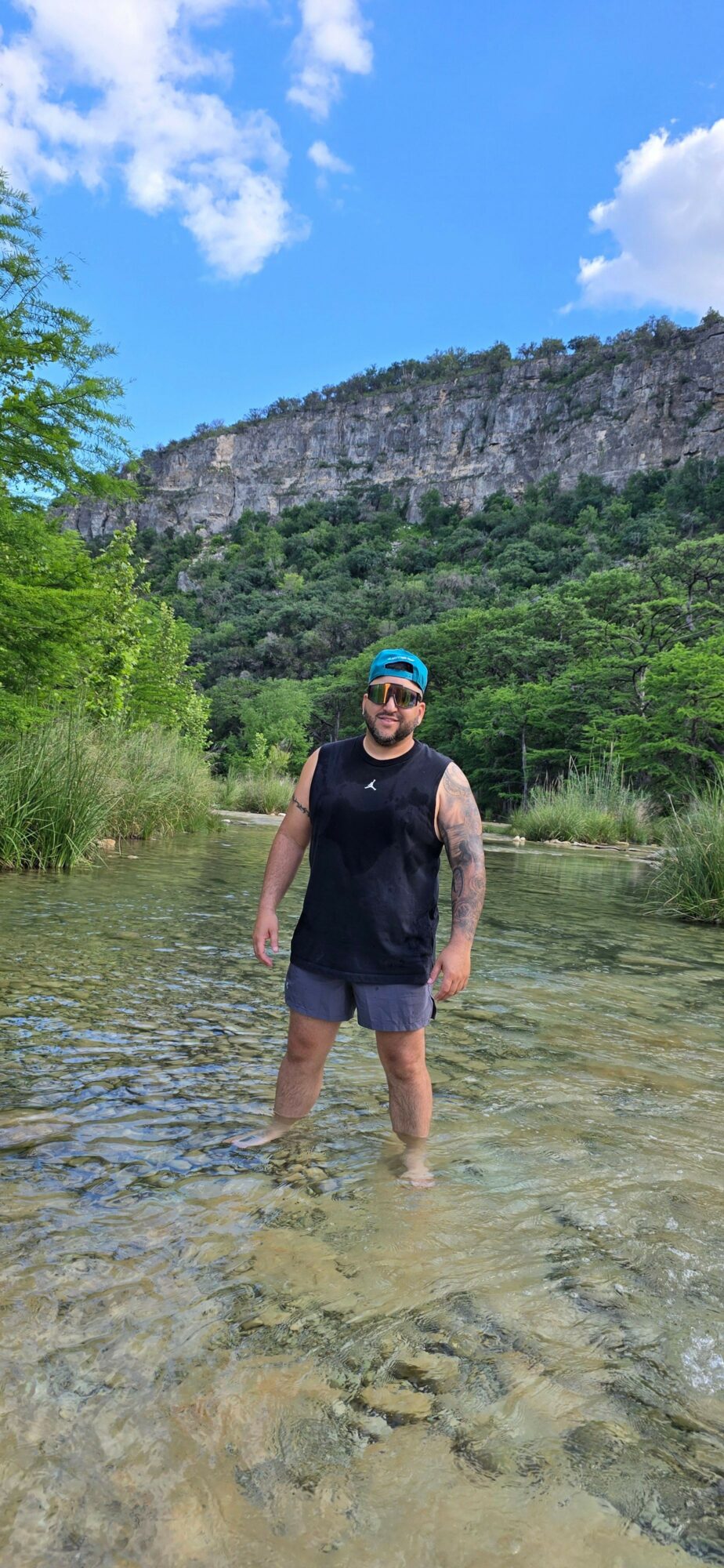 Person standing in shallow water with greenery and rocks in background, wearing a hat and sunglasses.