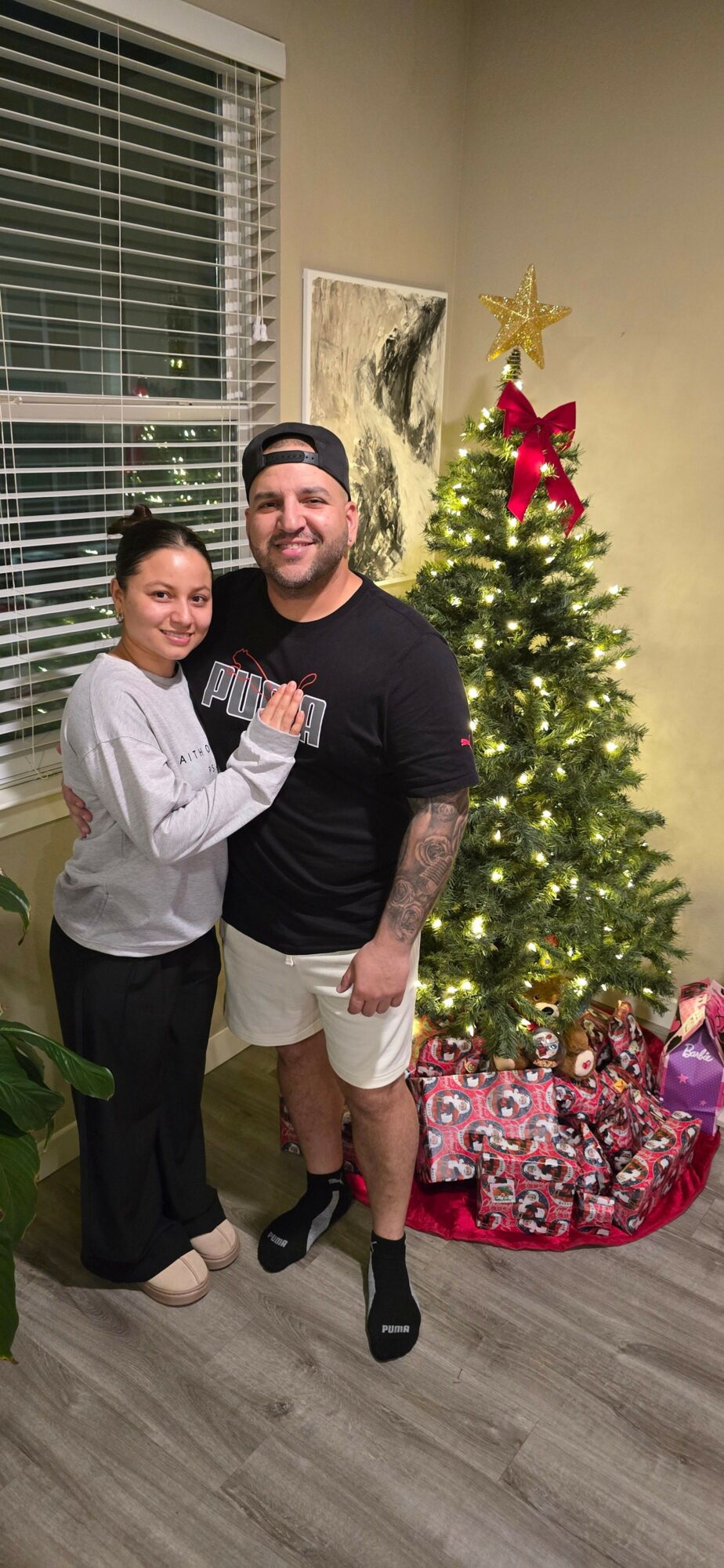 Two people standing in front of a decorated Christmas tree, smiling, indoors with a window and blinds behind them.