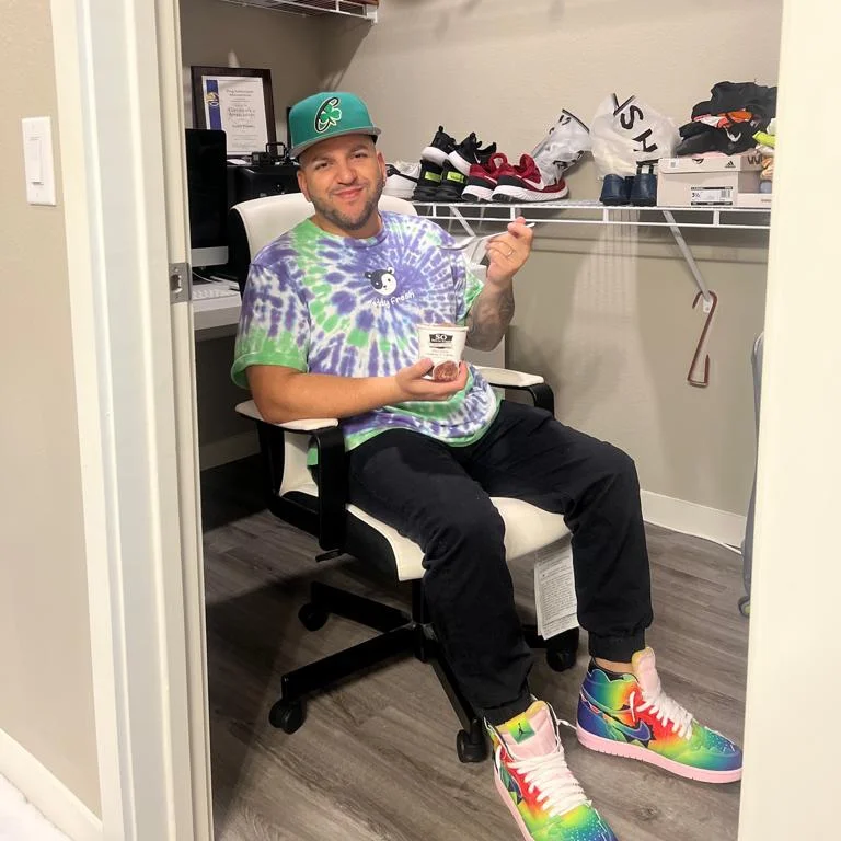 Person sitting in a chair holding a cup, wearing colorful tie-dye shirt and rainbow sneakers, in a closet with shoes and clothes.