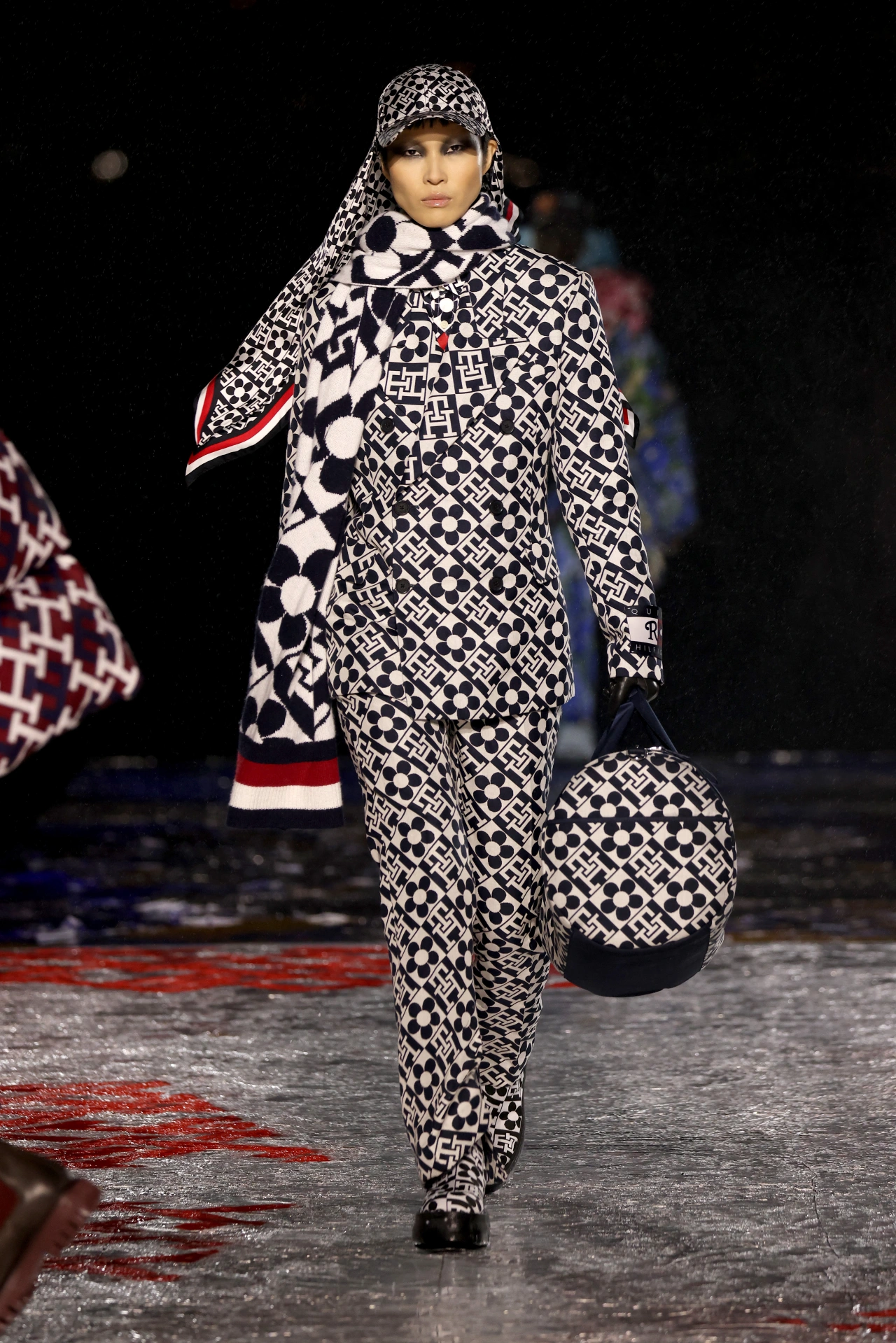 Model walking on runway wearing black and white patterned outfit, holding a matching helmet, with a scarf and hat, dark background.