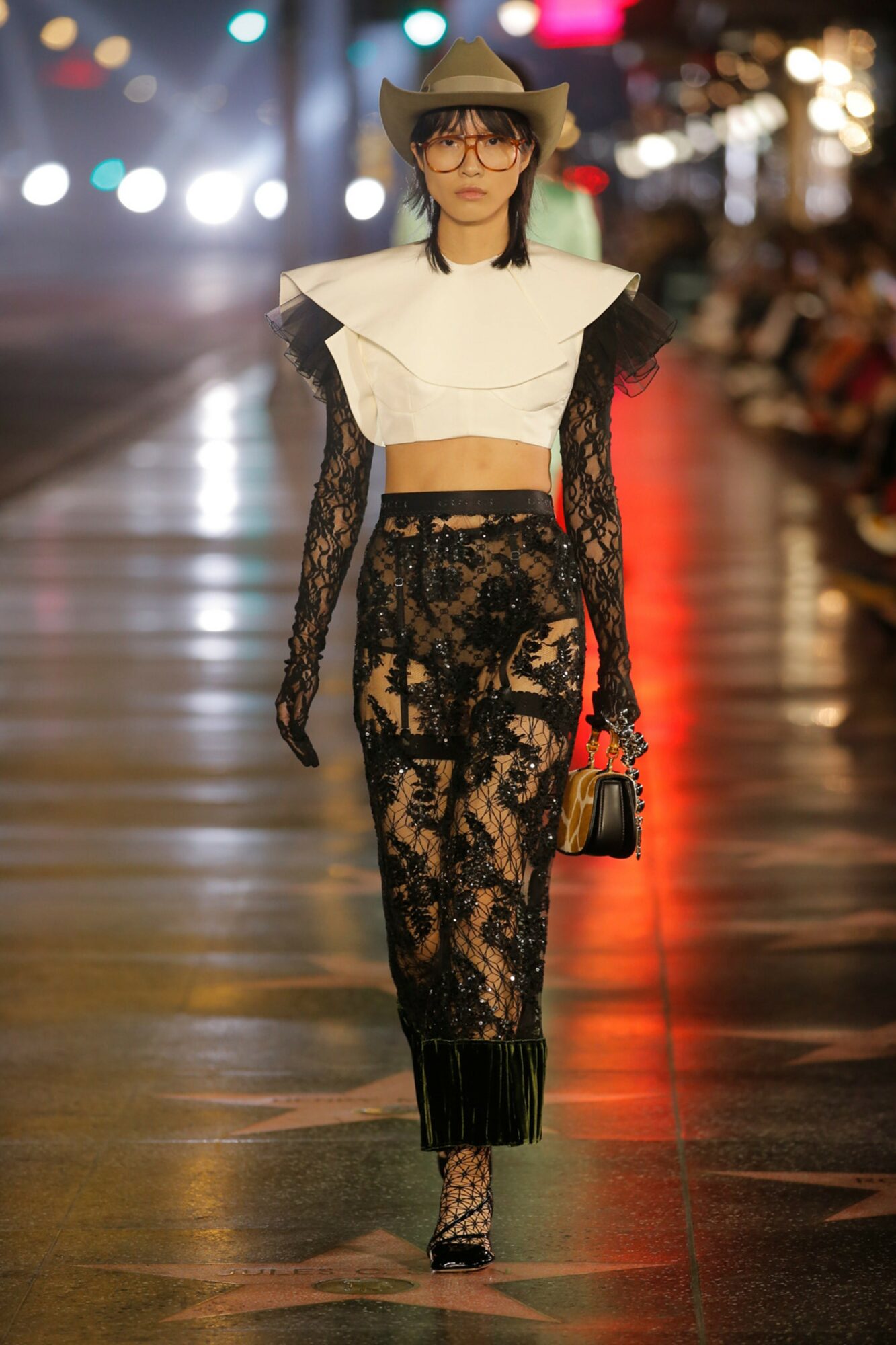 Female model walking on runway in black lace outfit with white collar, hat, and glasses, holding a small bag, nighttime setting.