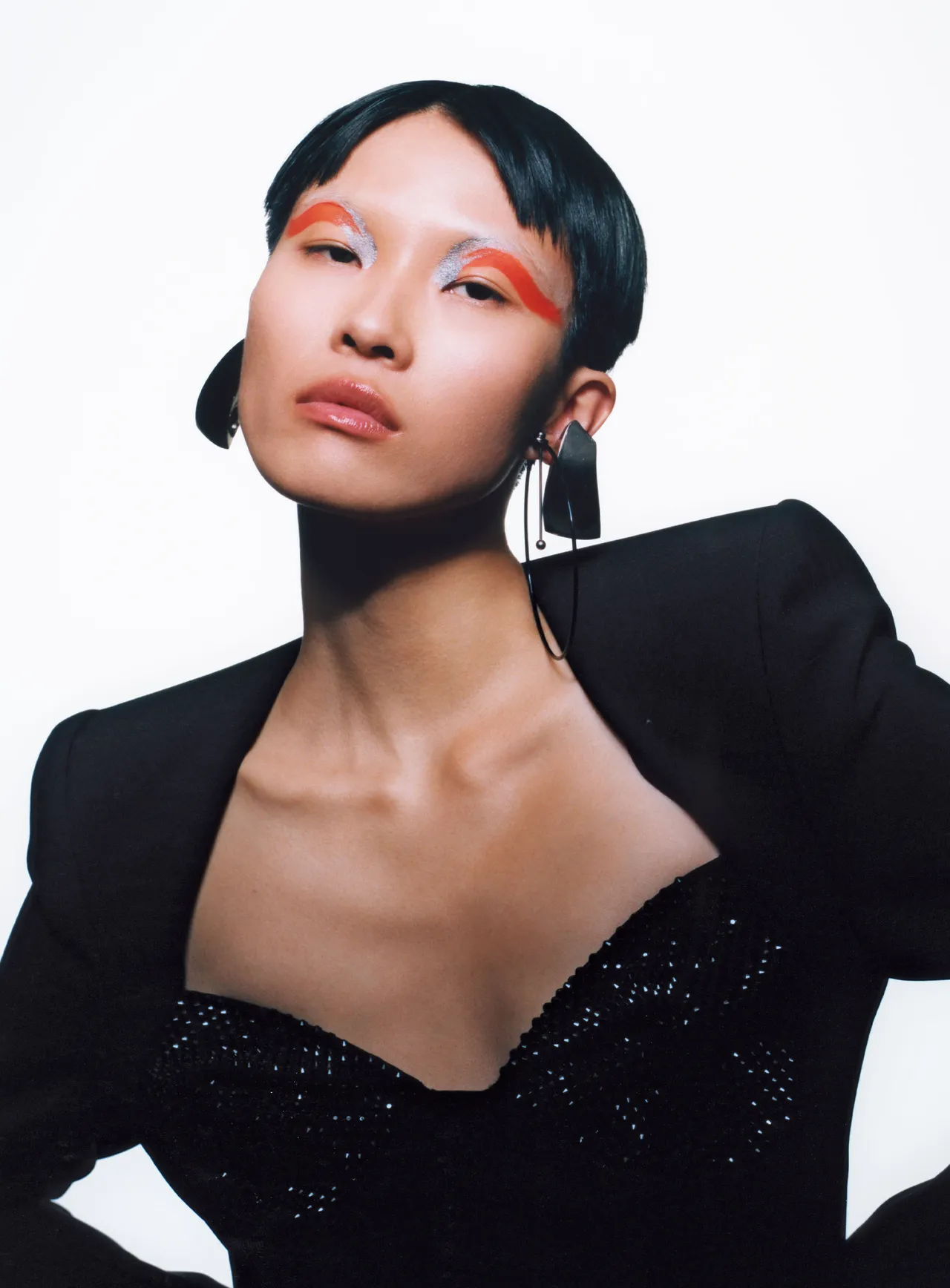 Woman with short black hair and colorful eye makeup, wearing a black outfit with a deep neckline, posing confidently against a white background.