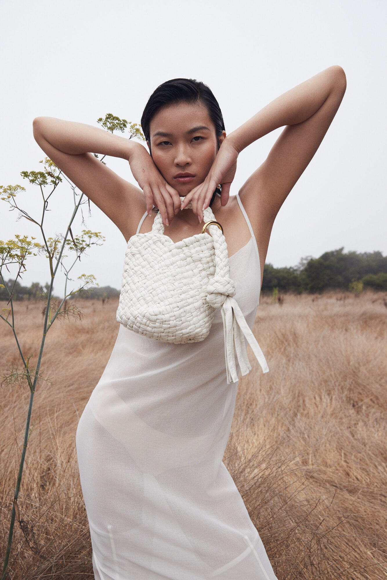 Woman in white dress holding a small woven bag outdoors, with arms raised and a serious expression.