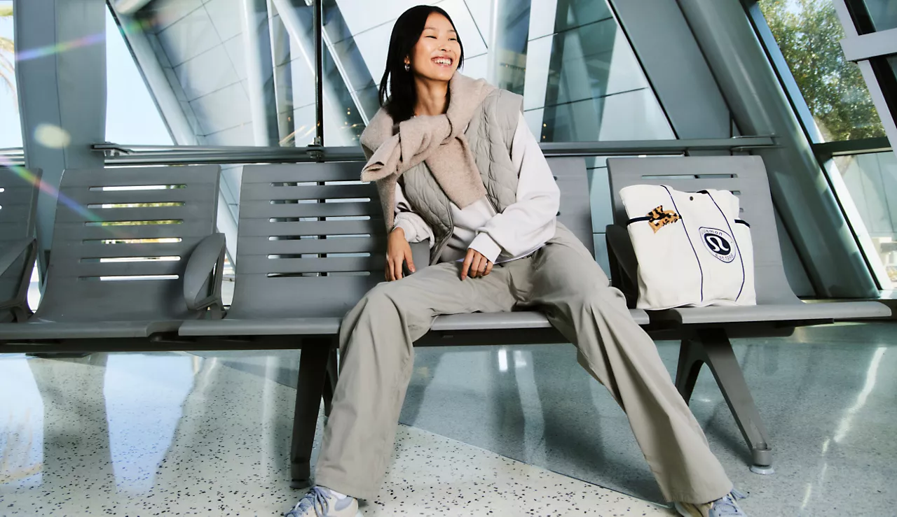 Woman sitting on a bench in an airport terminal, smiling, with a white bag beside her, glass windows behind her.