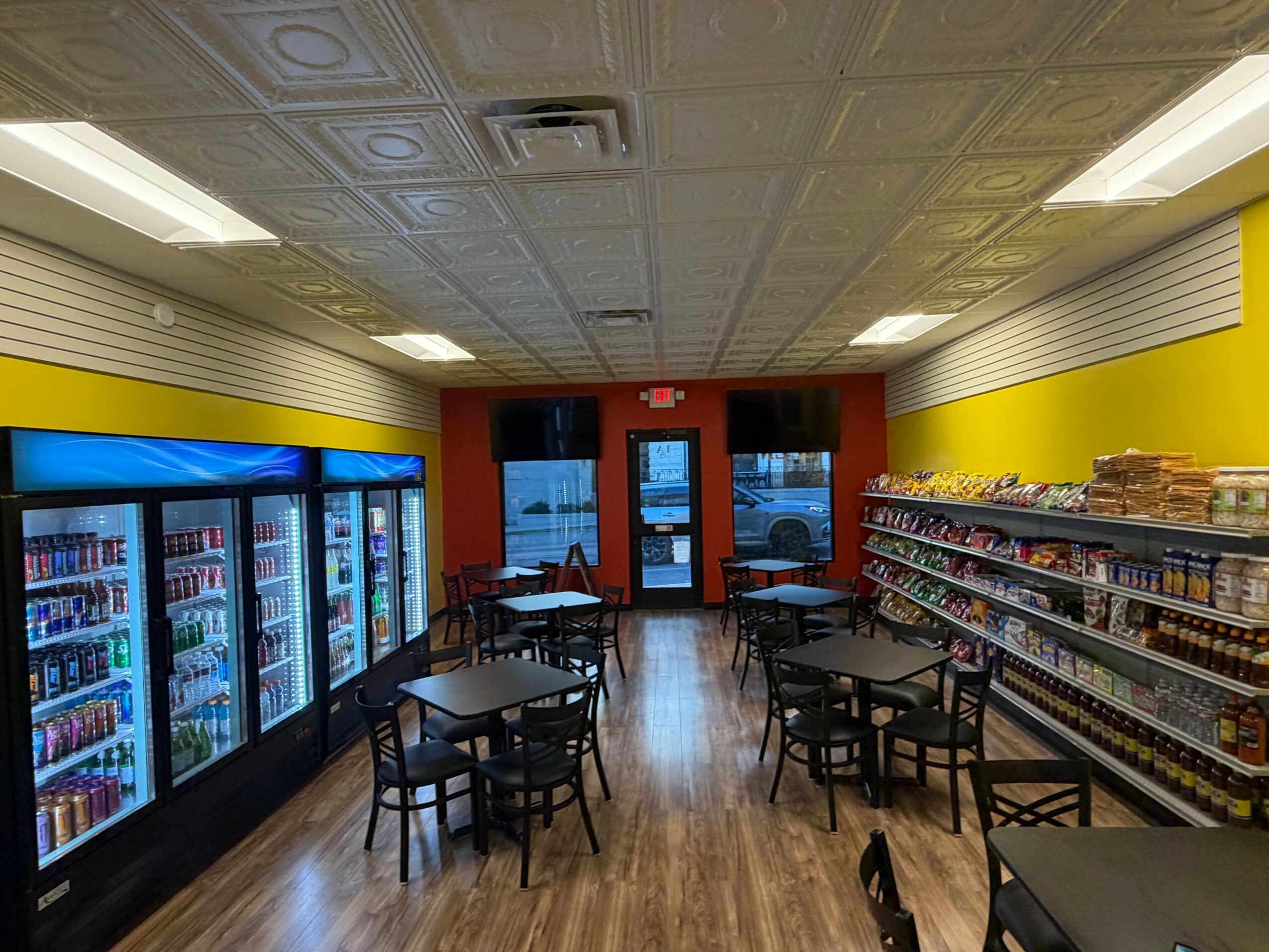Convenience store interior with refrigerated drinks, snack shelves, and seating area near glass doors, ceiling tiles, and bright lighting.