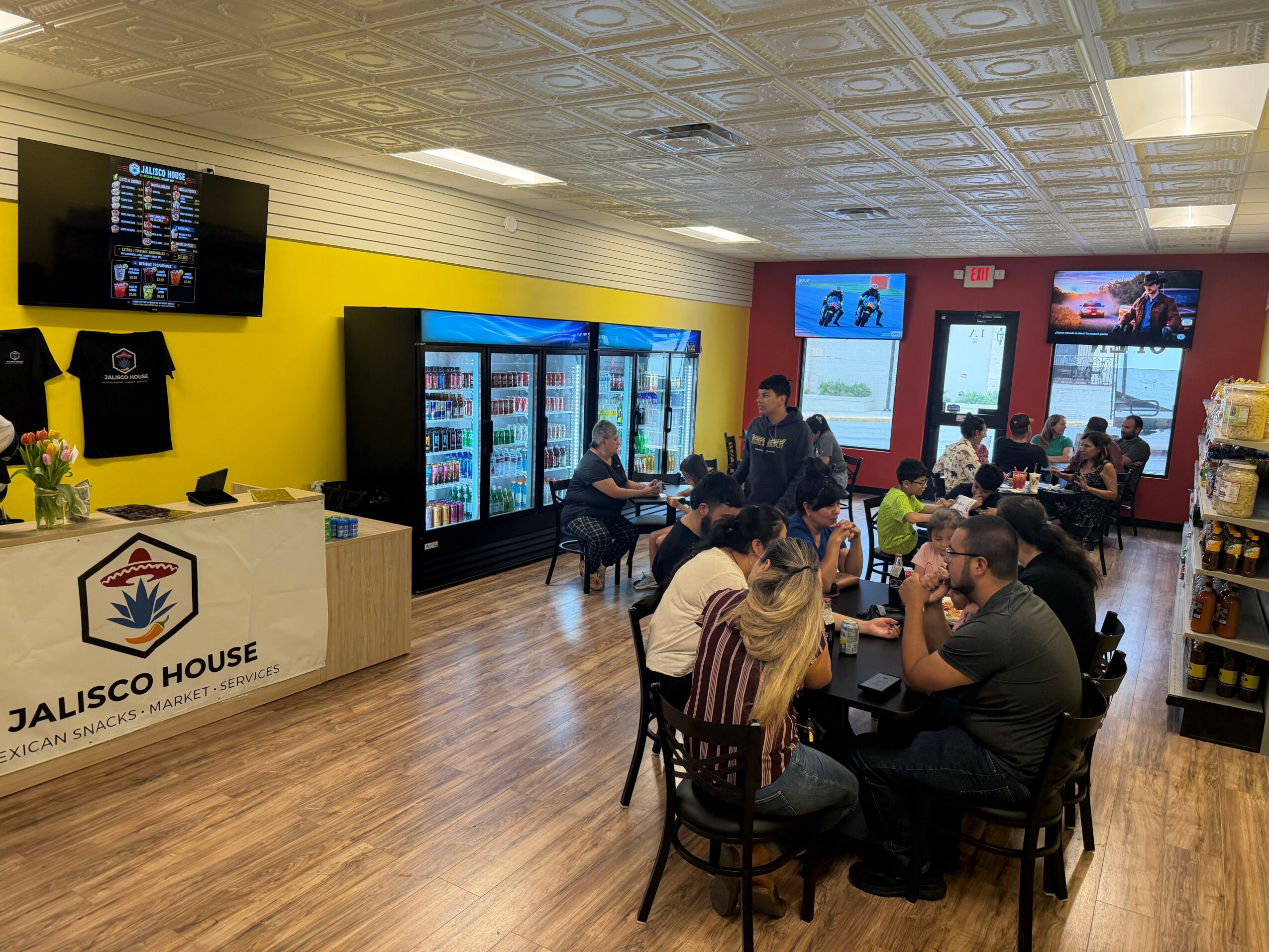 People sitting at tables in a restaurant with yellow and red walls, vending machines, and a TV screen, dining and socializing.