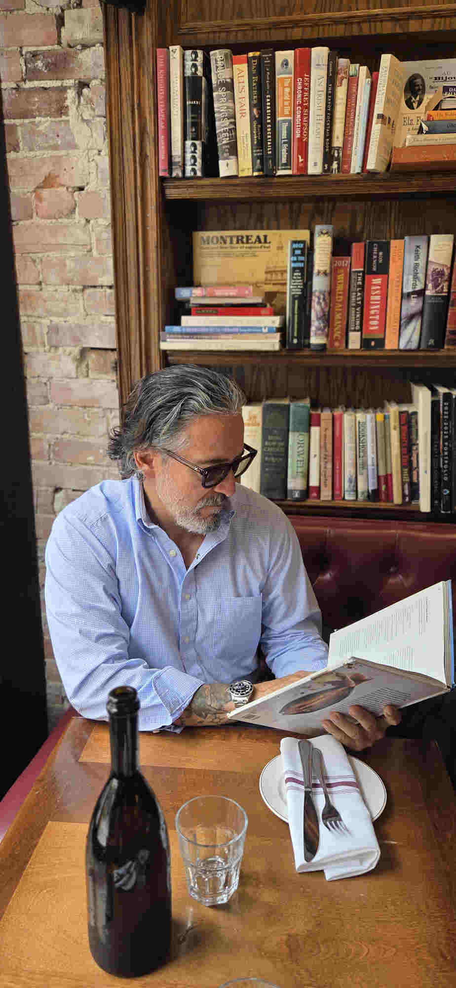 Man with gray hair and glasses reading a book at a table in front of a bookshelf, with a bottle and glass on the table.