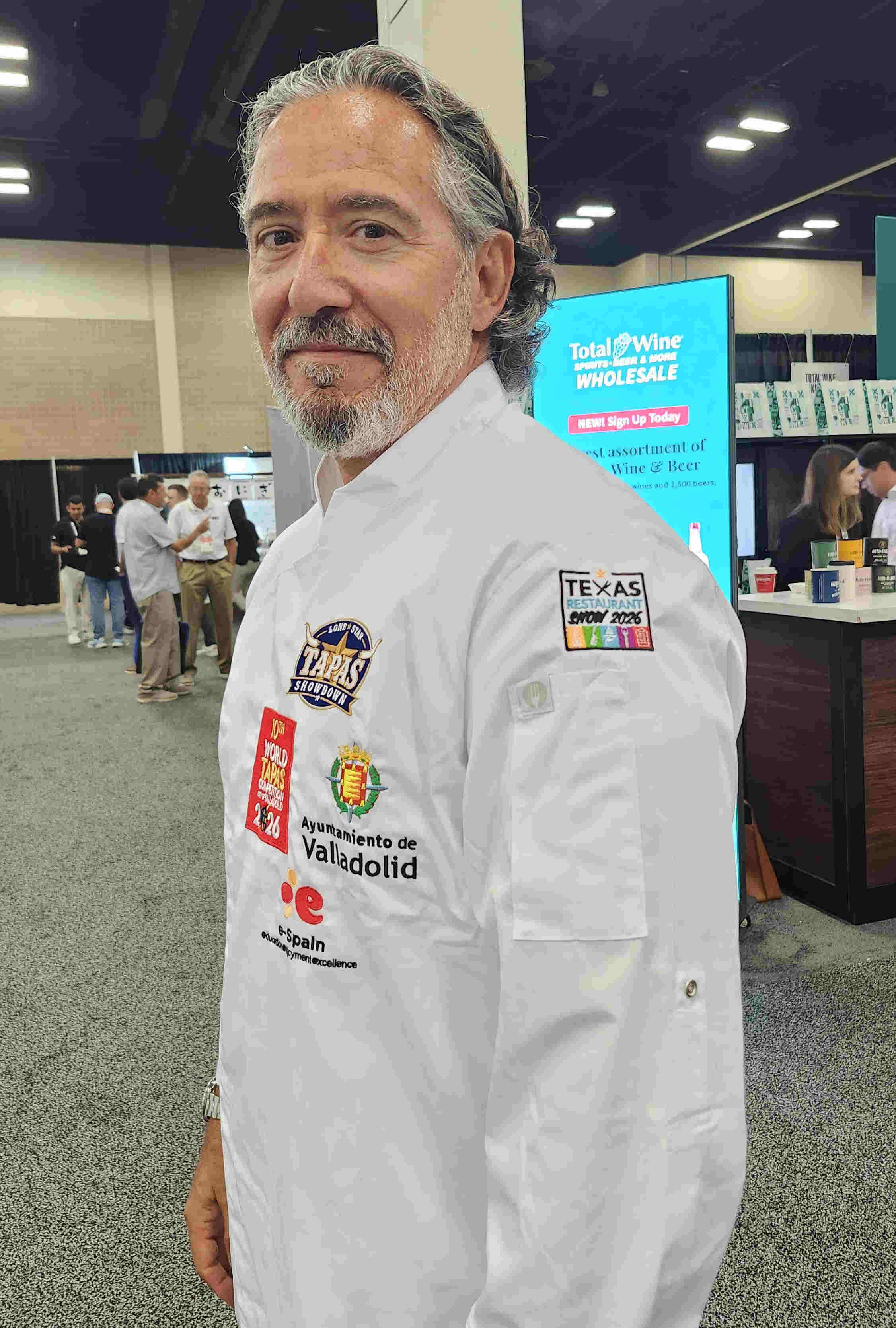 Man with gray hair and beard wearing a white chef's coat at an indoor event, with a digital screen and people in the background.