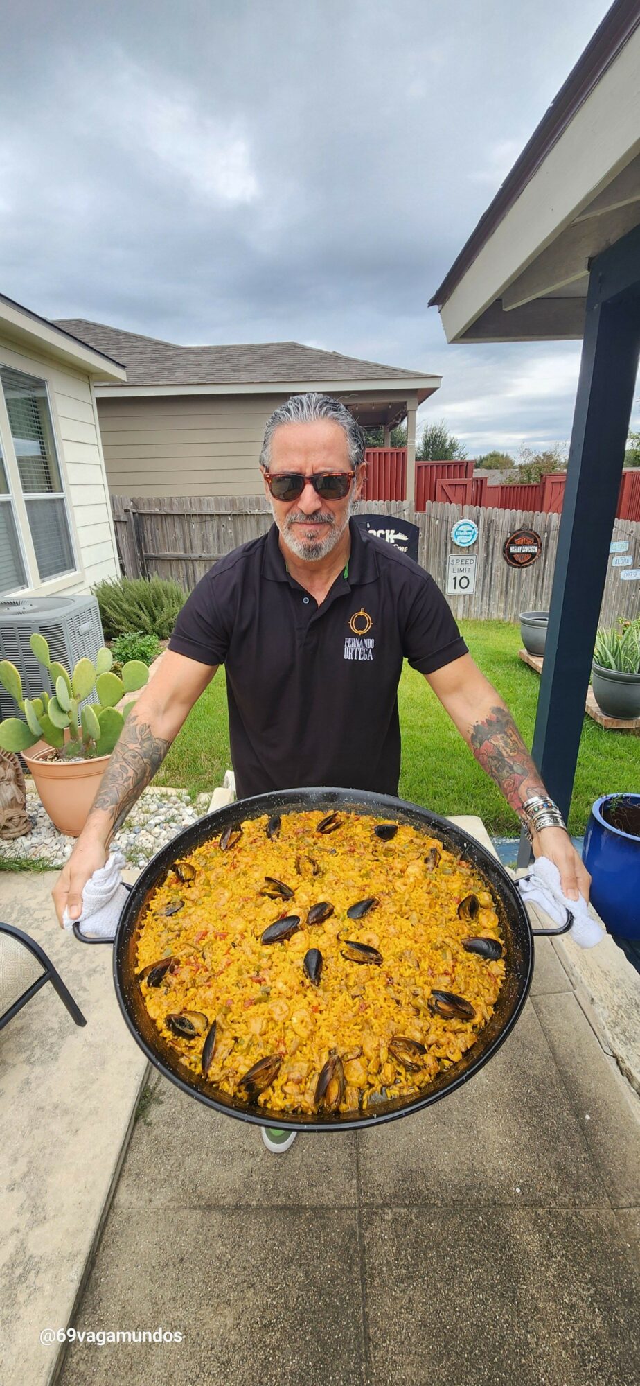 Person holding a large pan of paella outdoors on a patio, with a house and cloudy sky in background.