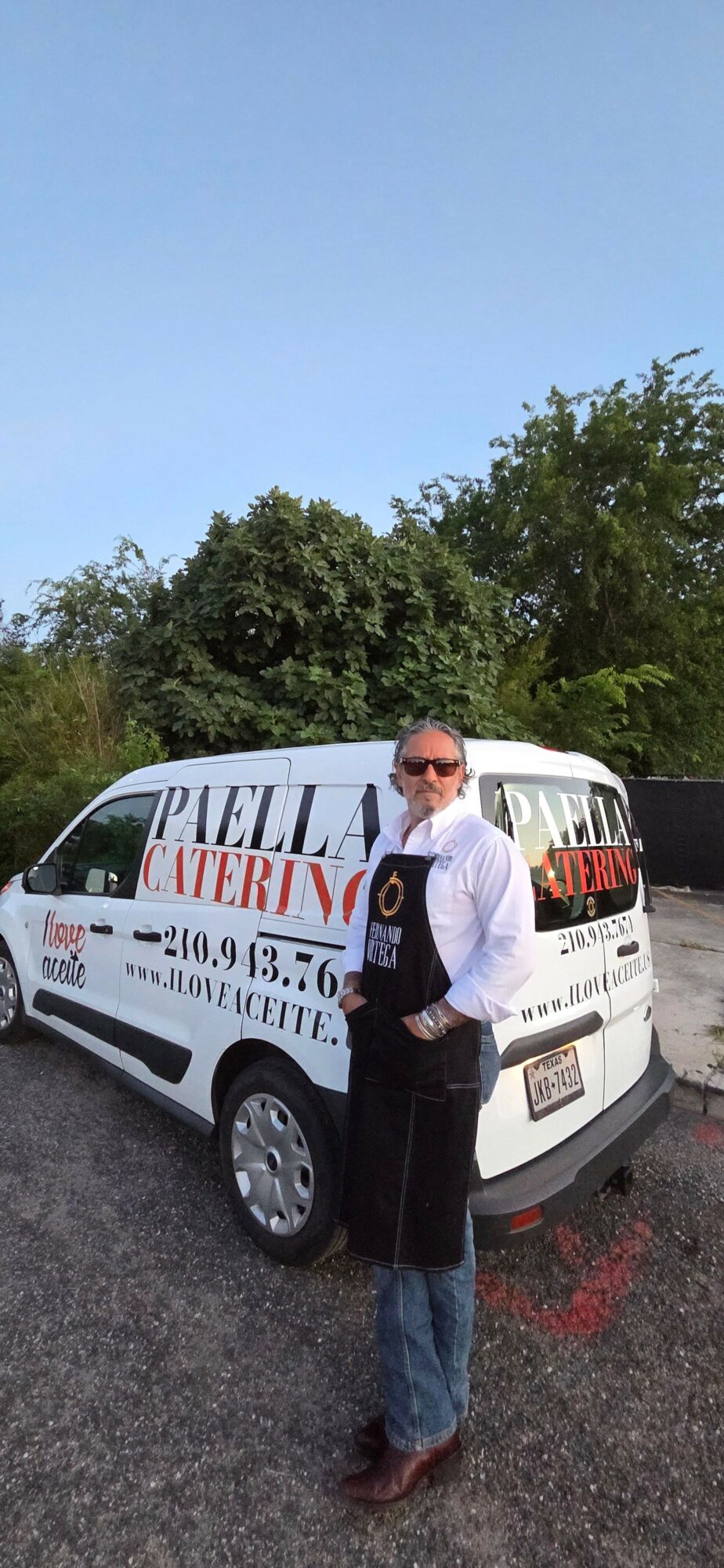 Person standing next to a white vehicle with advertising, outdoors on a paved surface, with trees and sky in background.