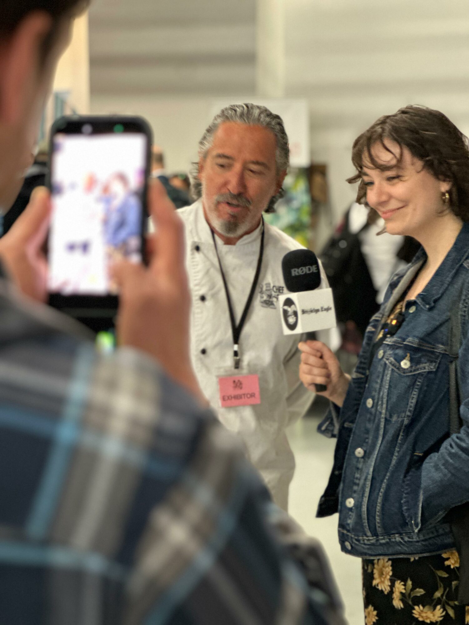 Person holding a phone records an interview with a woman and a man at an event, with a blurred background.