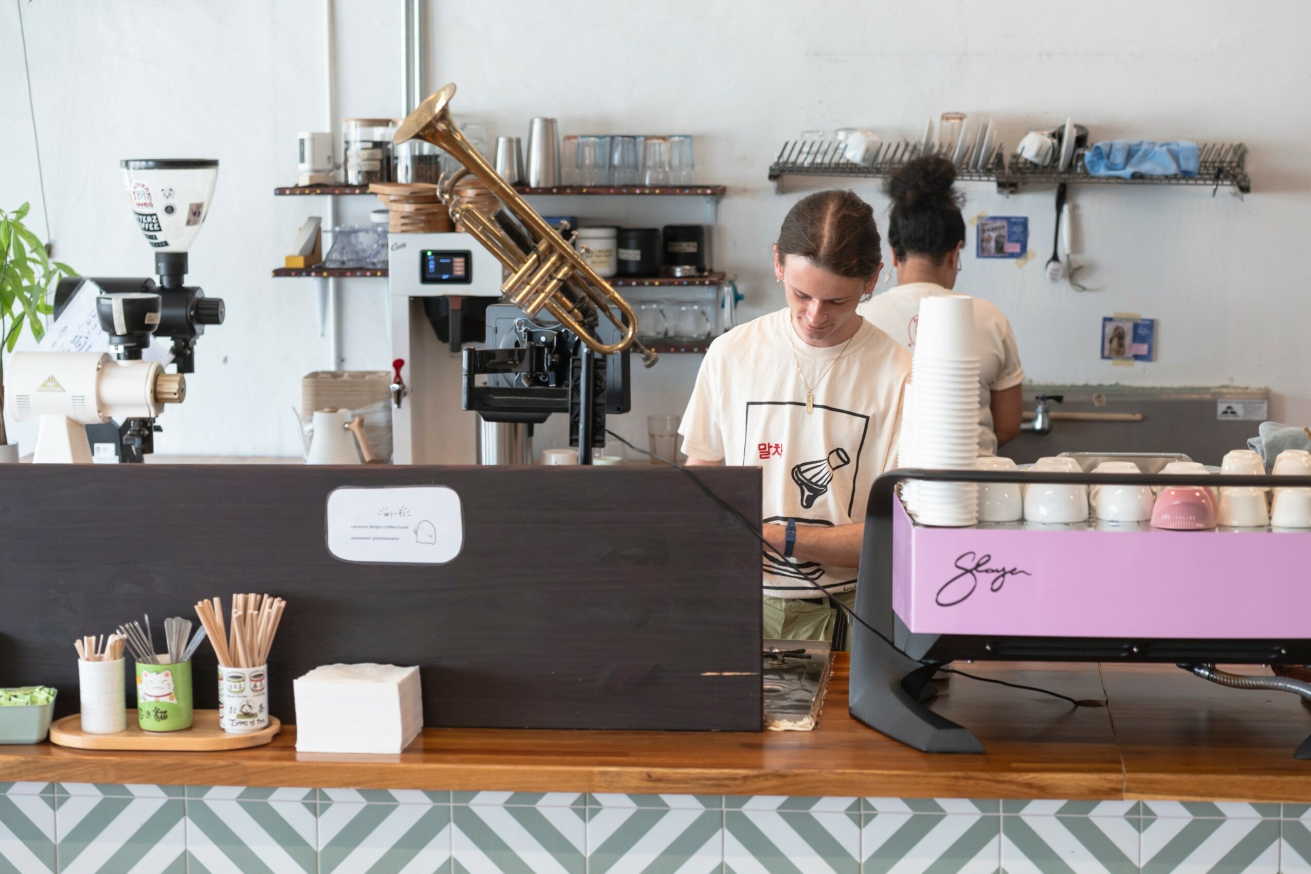 Two baristas work behind a coffee counter with equipment and supplies, one looking at a phone, the other with back turned.
