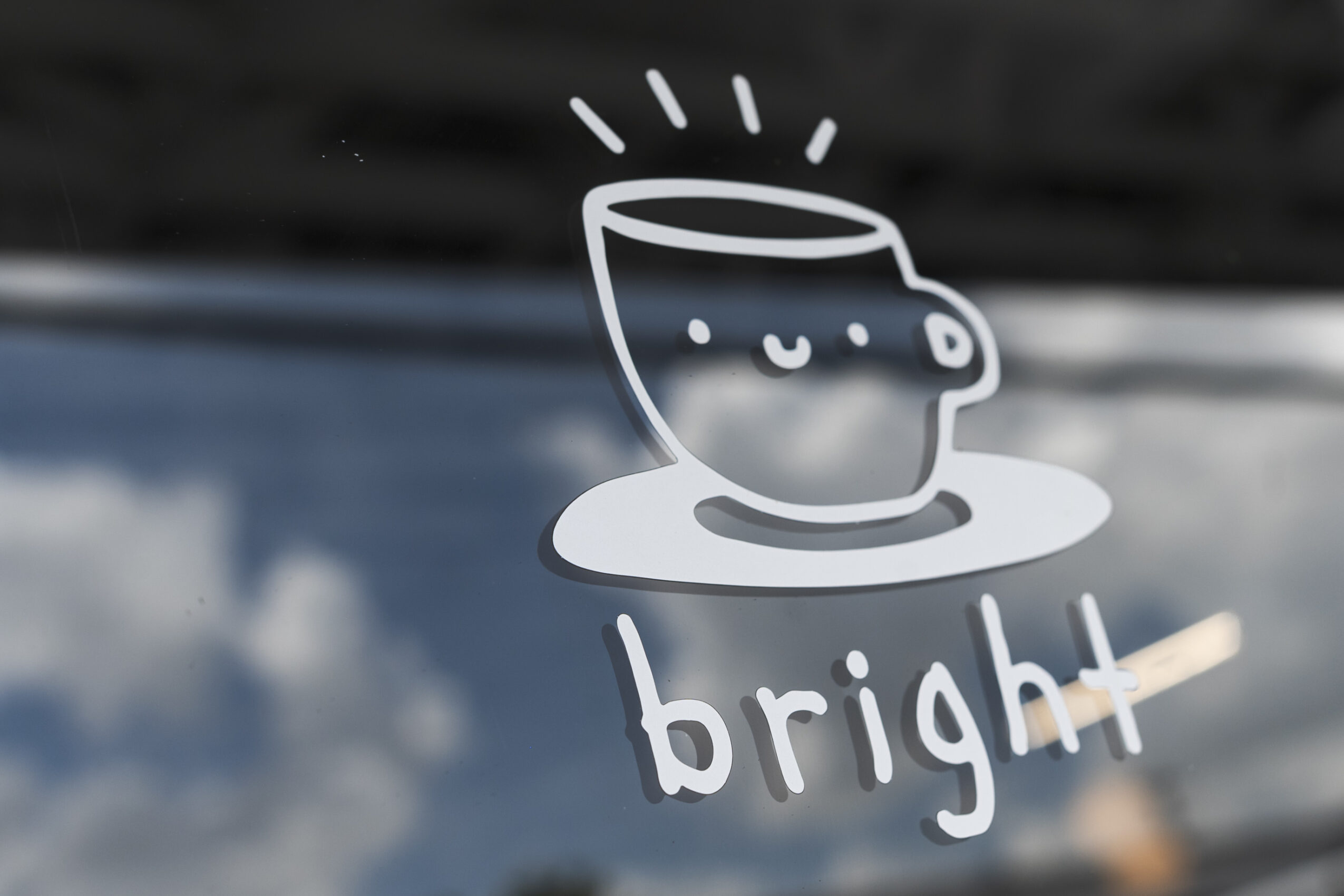 White line drawing of a smiling cup with steam above, and the word bright underneath, on a blurred background.