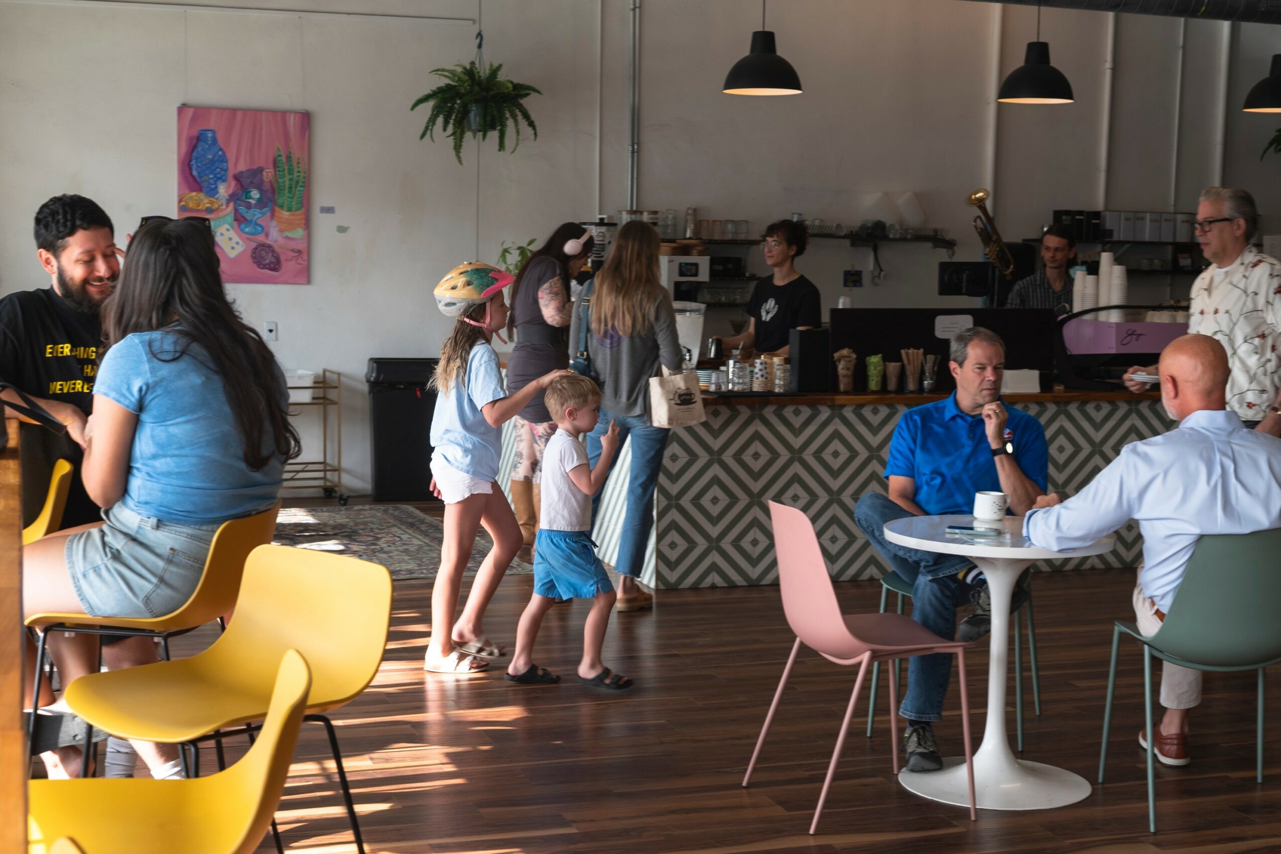 People gather in a cafe with children and adults, some sitting at tables and others standing, in a modern space with artwork and plants.