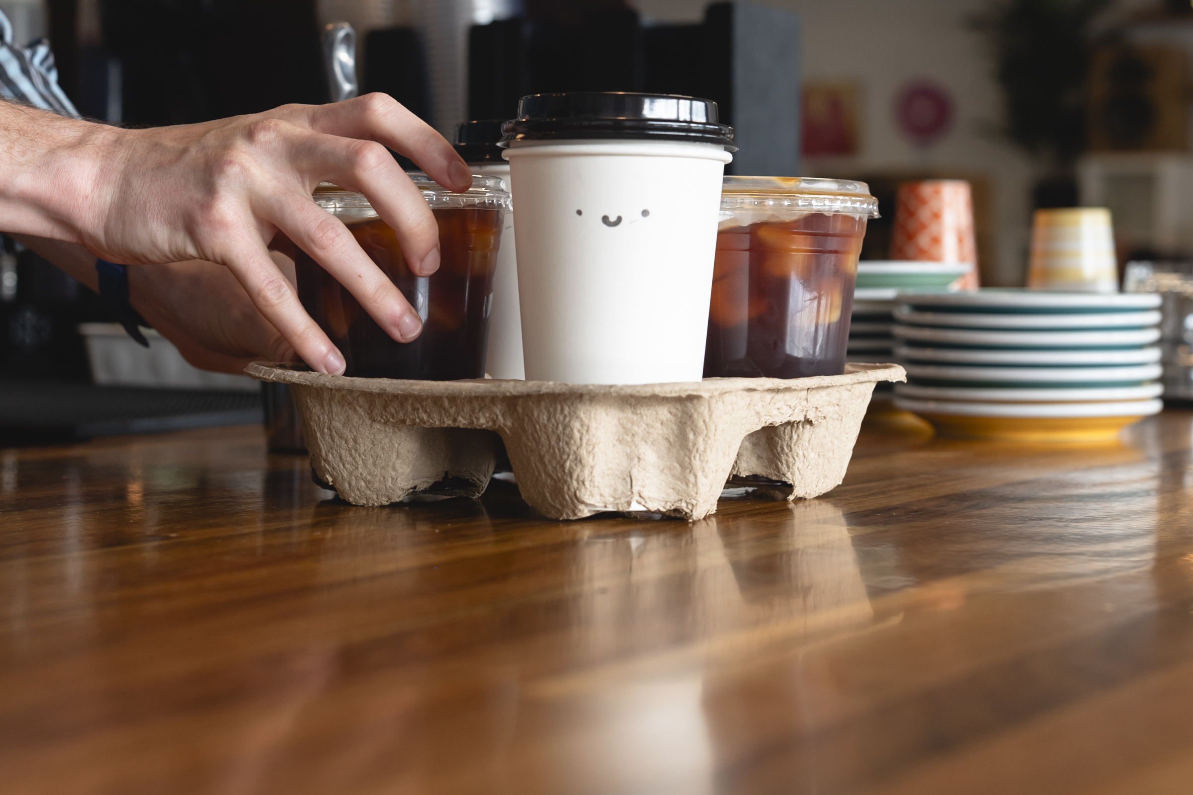 Hand reaching for a drink on a tray with coffee cups and plates on a wooden table.