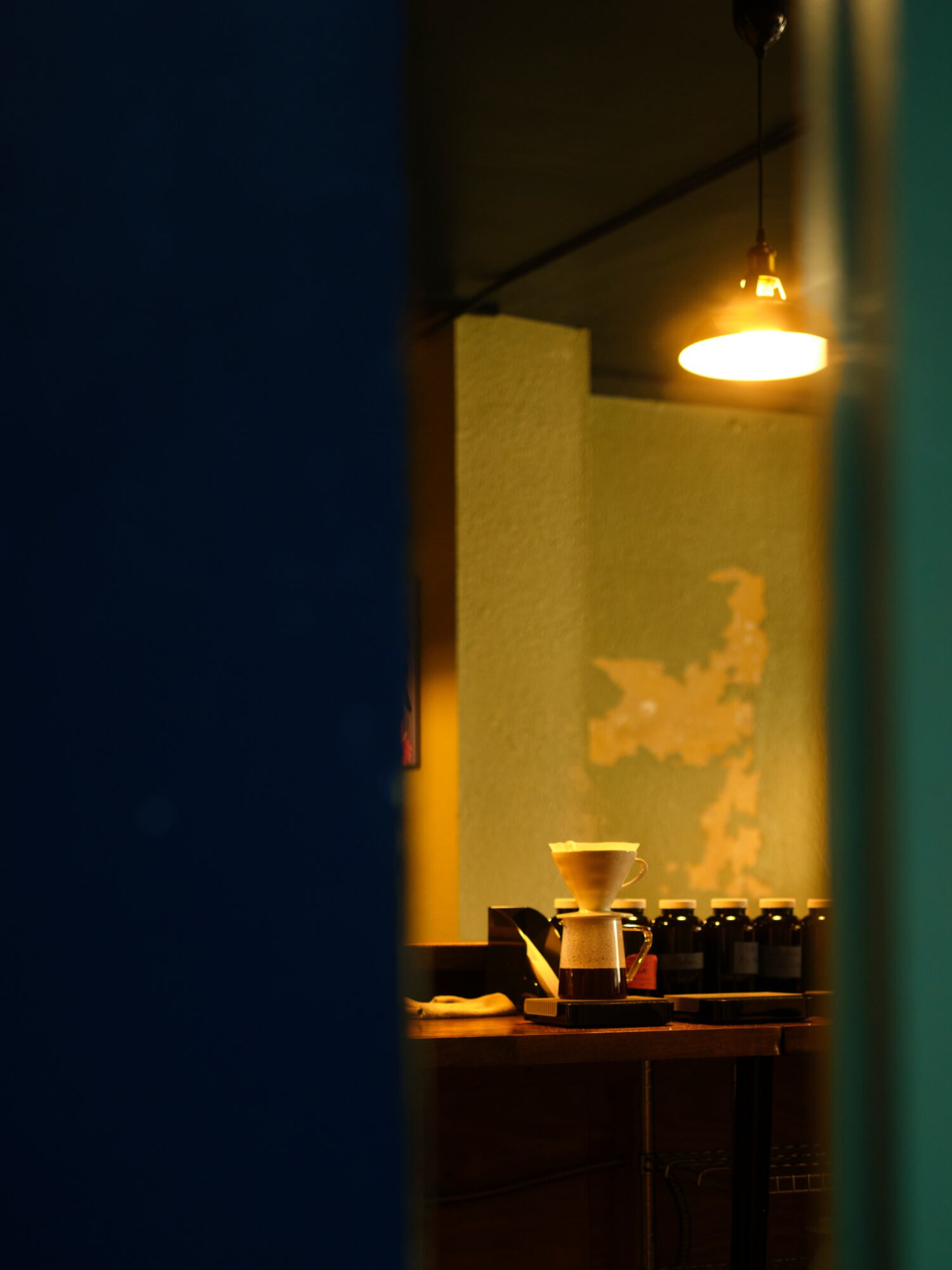 A dimly lit interior with a hanging light, a coffee cup, and bottles on a counter, viewed through a narrow opening.