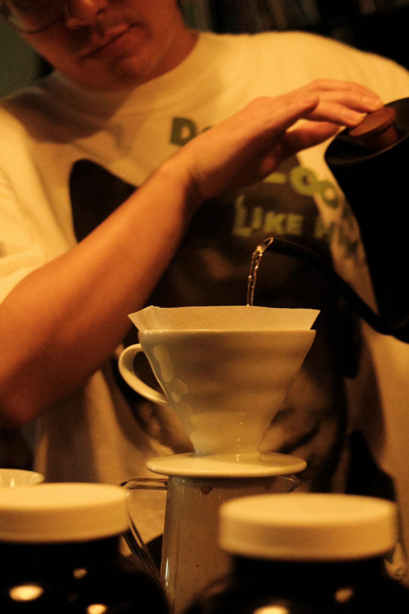 Person pouring hot water into a coffee dripper with cups and jars nearby.