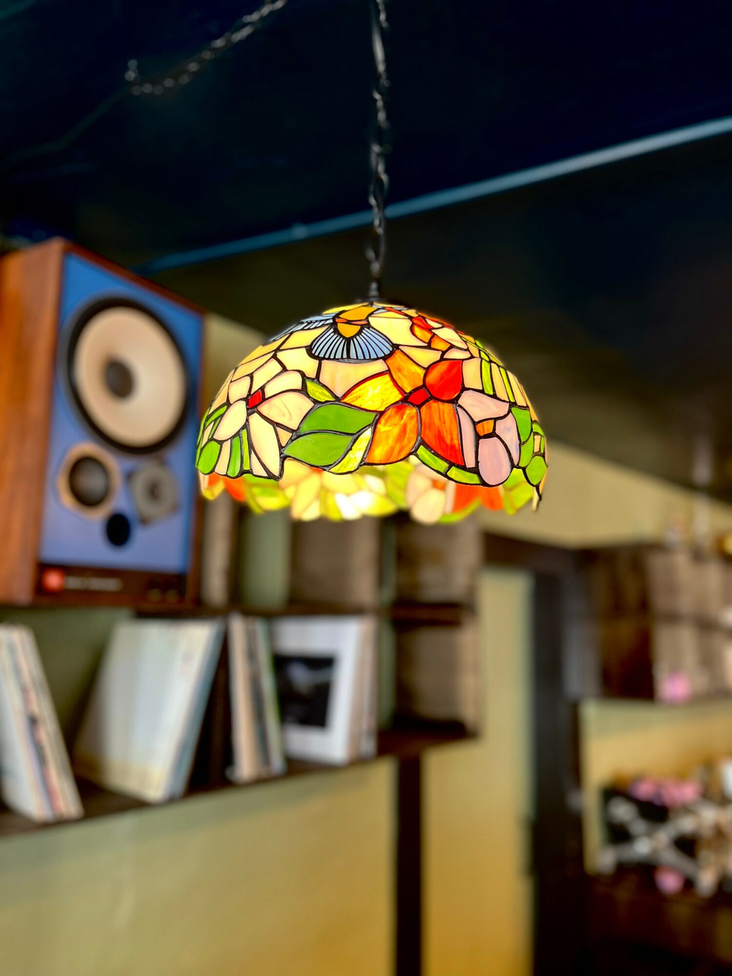 Colorful stained glass pendant lamp hanging from ceiling, with bookshelf and speaker in background.